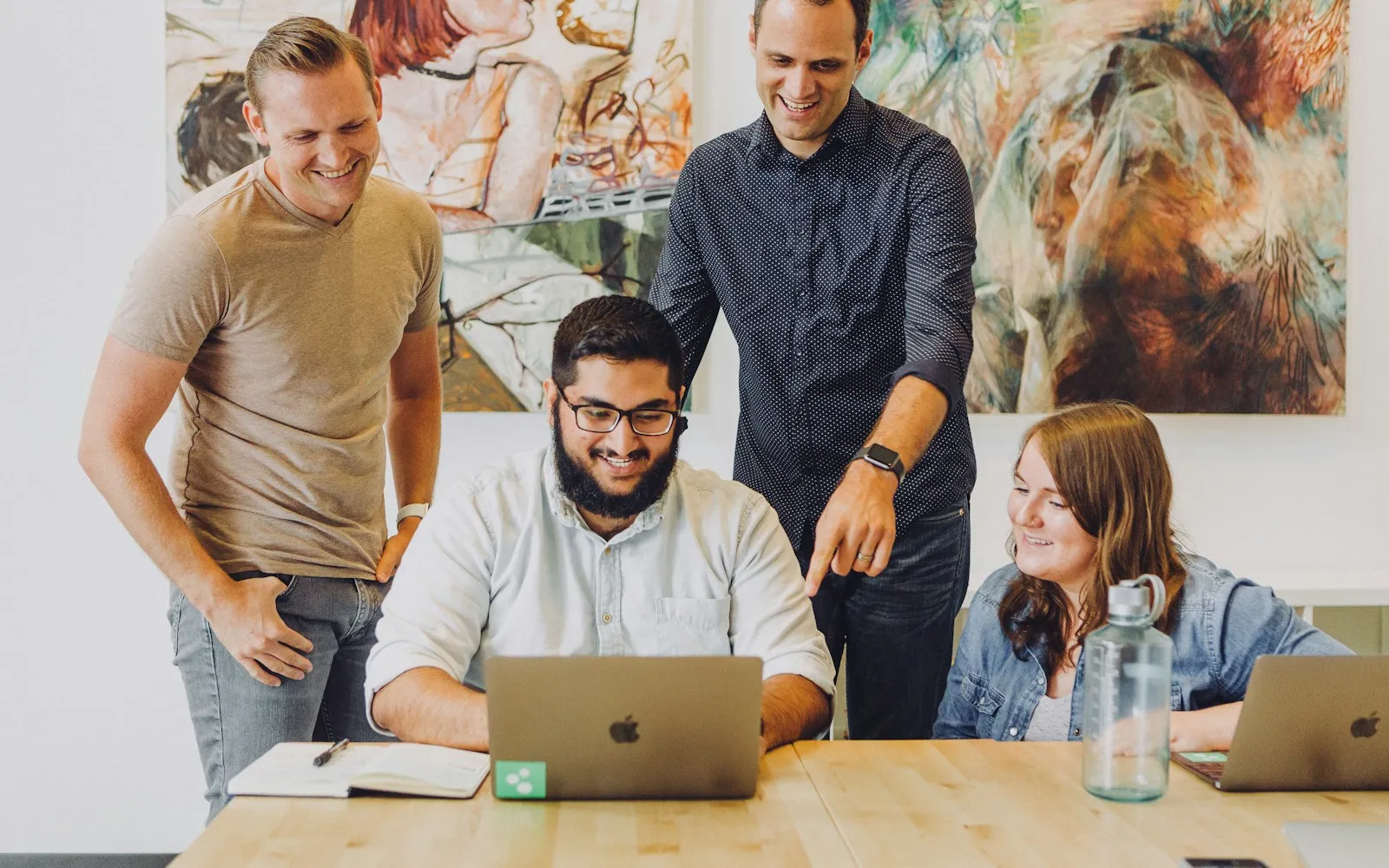 Four colleagues smiling around a laptop at a shared table.